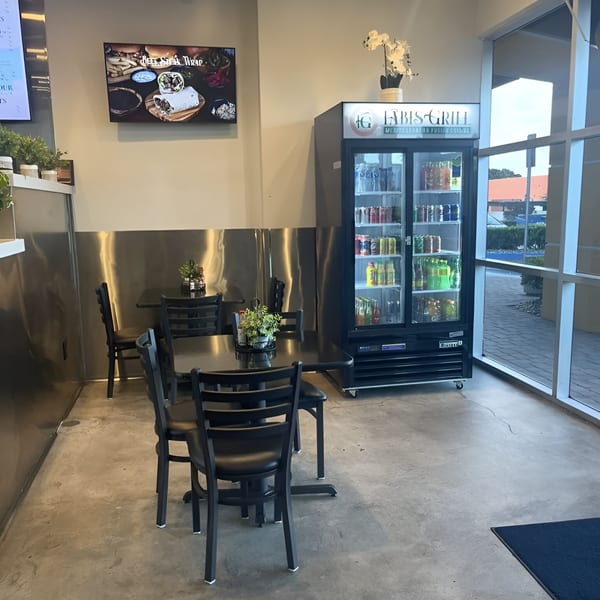 Interior of Fabi's Grill with black tables and chairs, a beverage cooler, and a TV displaying food.